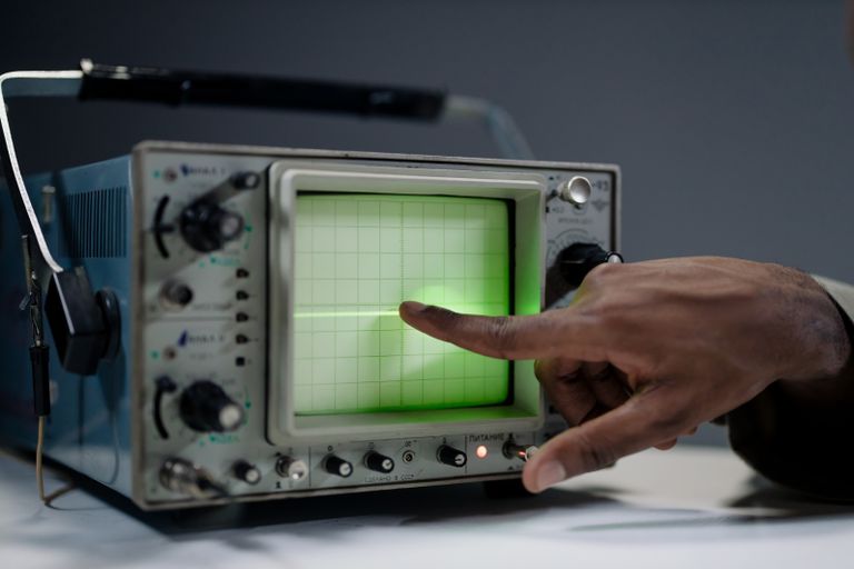 A hand pointing at the screen of a vintage oscilloscope indoors.
